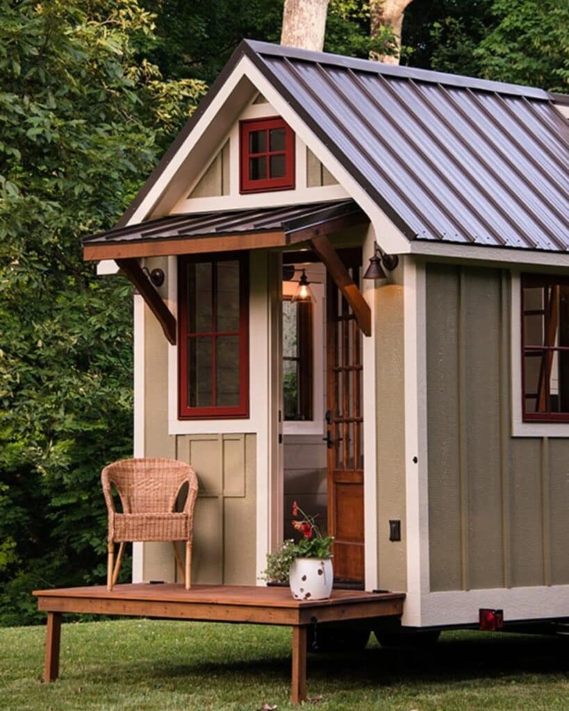 Exterior close-up of the Ynez tiny house entrance featuring a wooden porch, a wicker chair, and red-trimmed windows against tan siding.