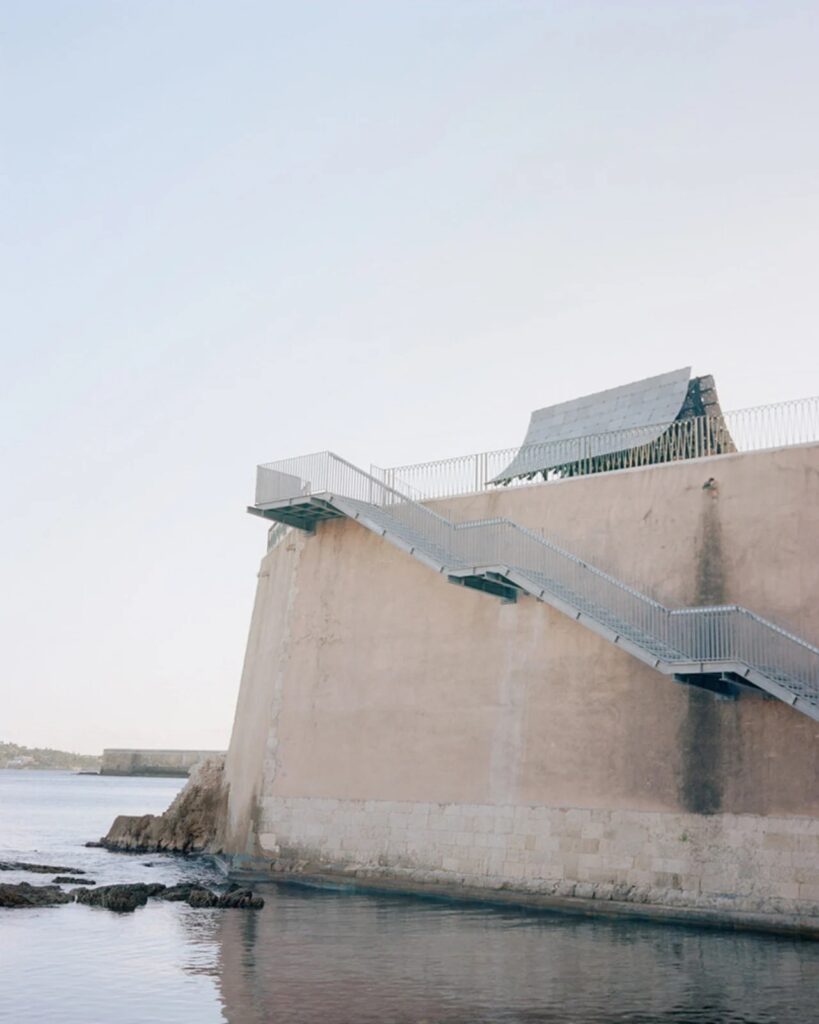 The Asympta structure seen from below a high stone fortification wall with a metal staircase leading up from the sea.