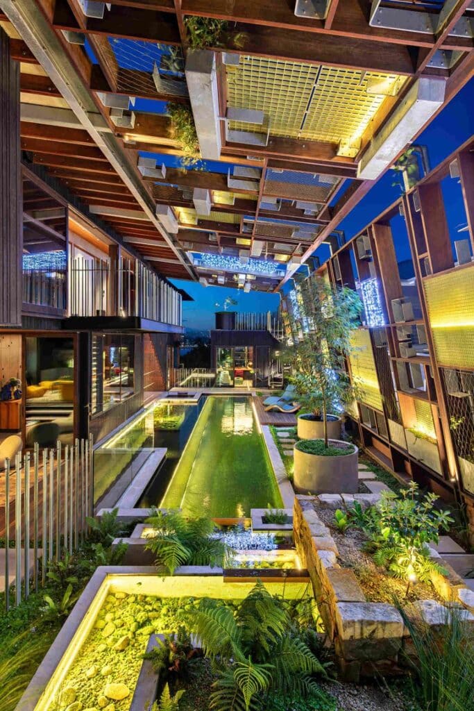 Night view of Holocene House featuring an illuminated natural pool, garden beds, and a complex overhead timber structure.