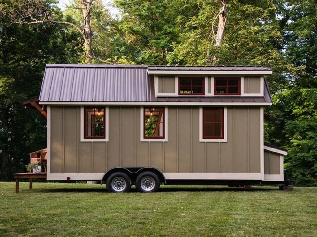 Full side profile of the Ynez tiny house on a double-axle trailer, featuring olive-green vertical siding, white trim, and a gabled metal roof.