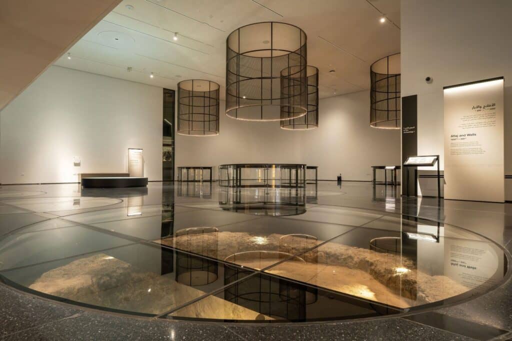 Modern museum gallery with glass floors revealing ancient wells and suspended cylindrical mesh light fixtures.