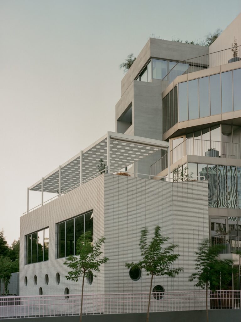 Close-up of a white brick building section with circular porthole windows and a rooftop terrace with a perforated metal canopy.