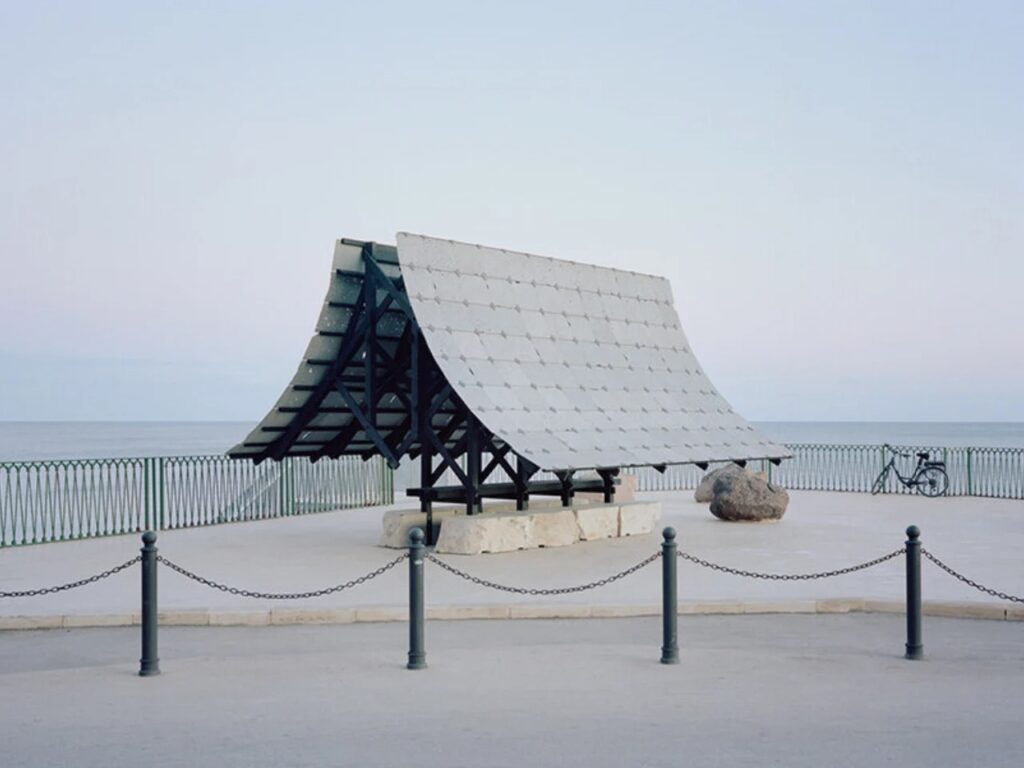 Full view of the Asympta installation on a waterfront plaza with a bicycle parked in the distance under a pale sky.