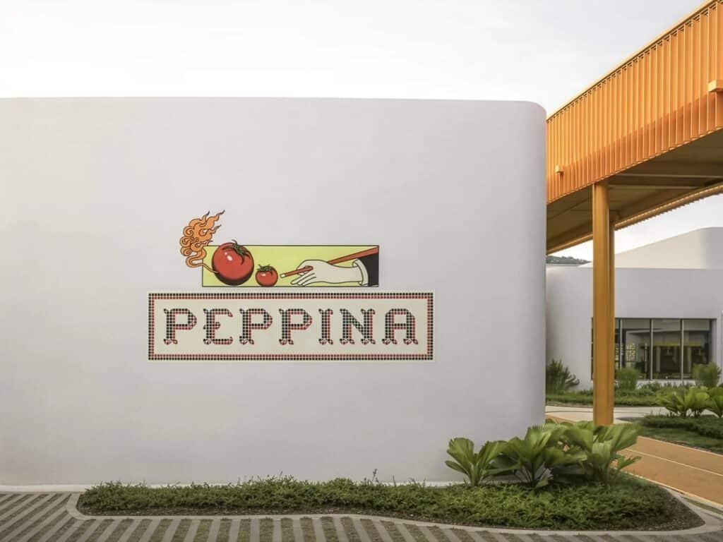 Exterior white curved wall of a retail building featuring a mosaic logo for Peppina restaurant, surrounded by tropical greenery.