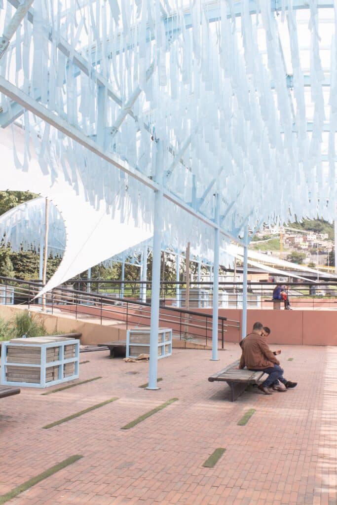 Two people sitting on a wooden bench inside the pavilion under a canopy of blue fabric strips, with an urban hillside visible in the distance.