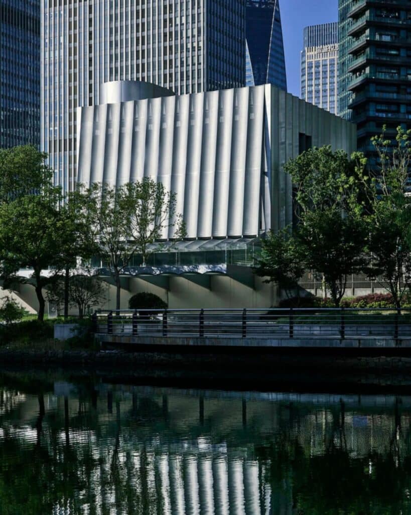 External view of Hangzhou Empathy Museum featuring undulating metallic facade reflecting on the water at daytime.