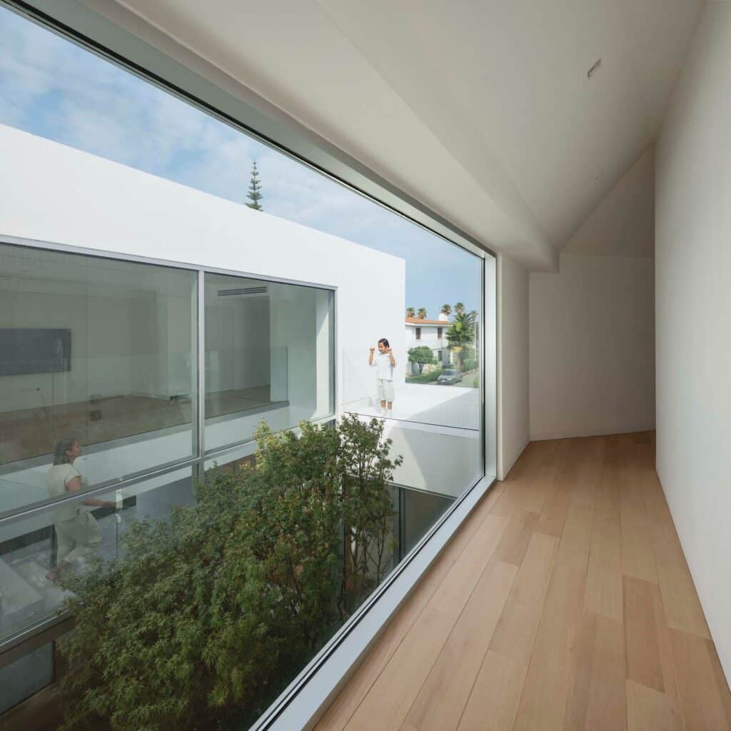 A bright interior corridor with wooden flooring and a large window overlooking the central tree and another part of the villa.