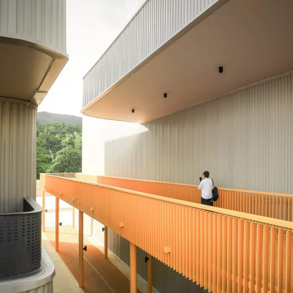 An elevated orange pedestrian walkway connecting modern corrugated metal buildings with a view of lush green mountains in the background.