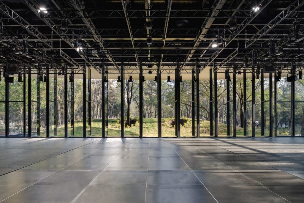 Interior view of a large multi-purpose hall with a professional lighting grid and glass walls overlooking the park.