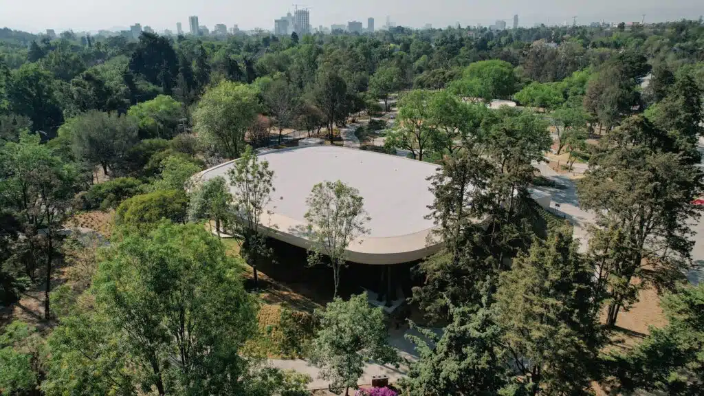 Aerial drone view of a large pavilion with a light-colored roof surrounded by a dense forest in Chapultepec Park.