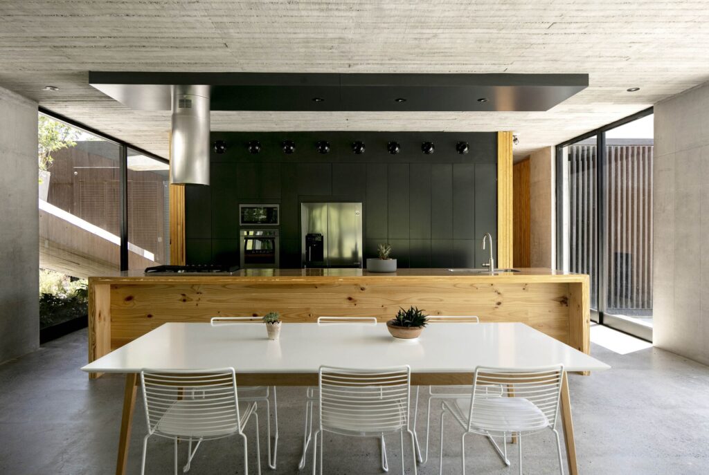 A modern interior featuring a long wooden kitchen island, white dining table, and black minimalist cabinetry against a concrete ceiling.