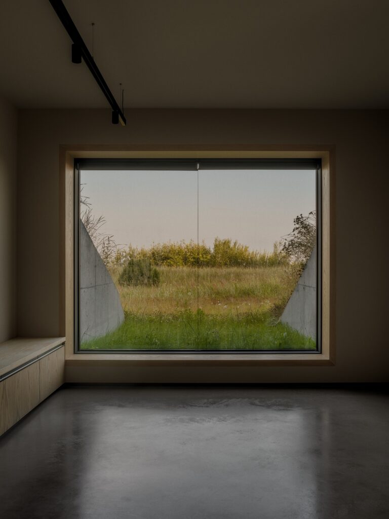 View through a large square window from a minimalist interior looking out at concrete walls and a grassy slope.