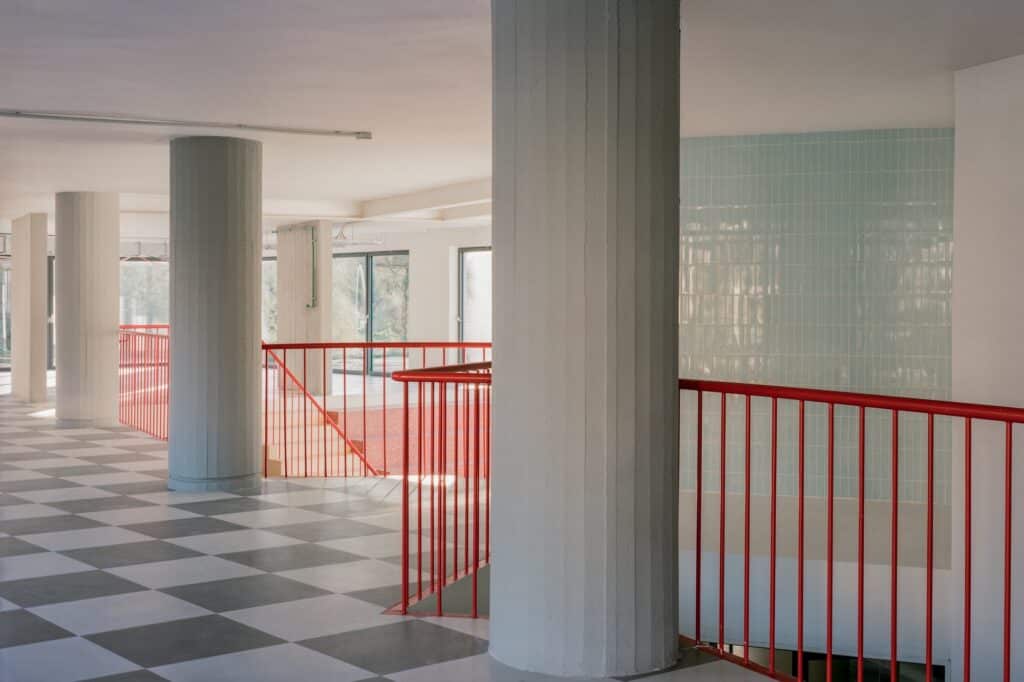 Internal hallway featuring a checkered floor pattern, massive concrete pillars, and bright red safety railings against a light blue tiled wall.
