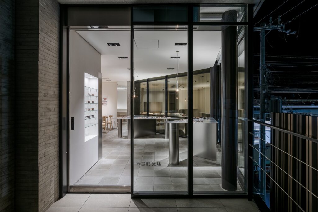 Exterior evening view of Ashiya Optical Store's glass entrance showing the internal layout and lighting.