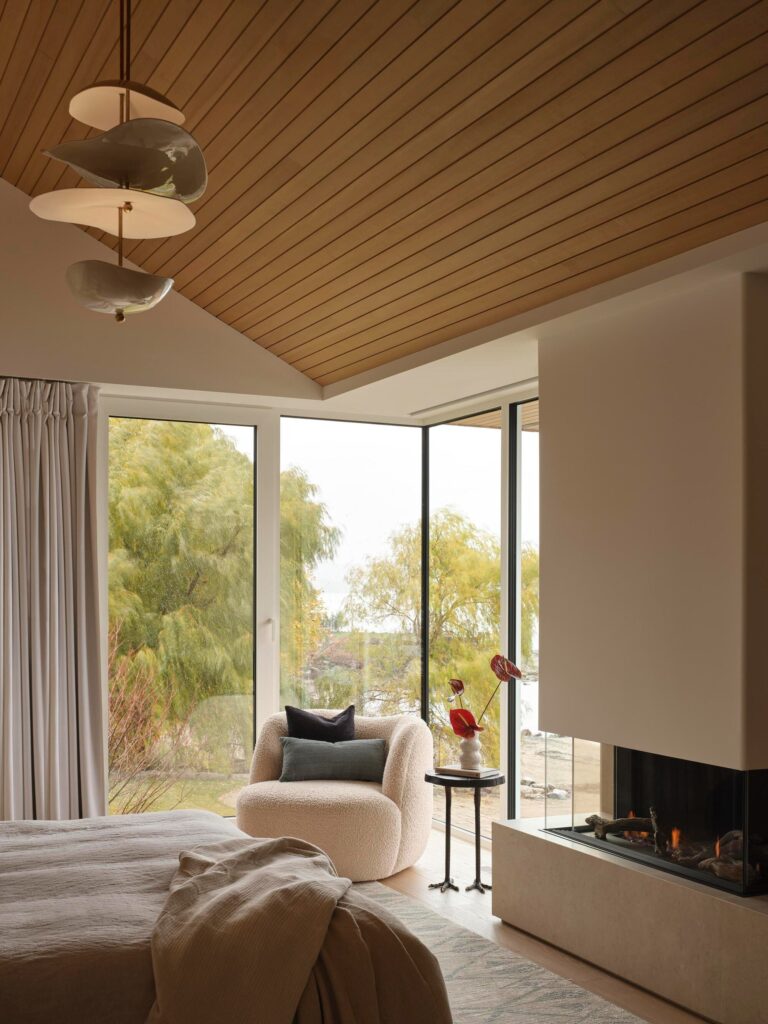 Master bedroom featuring a modern corner fireplace, cozy armchair, and large windows facing the coastal trees and shoreline.