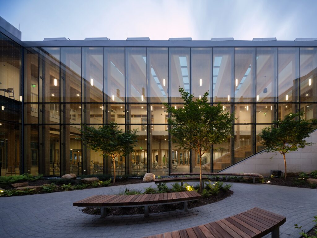 Ground-level view of an outdoor courtyard with curved benches and a glass wall reflecting the interior mass timber structure.