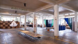 An industrial warehouse space transformed into an art exhibition with various installations, including a long stone table, hanging white fabric, and blue barrels.