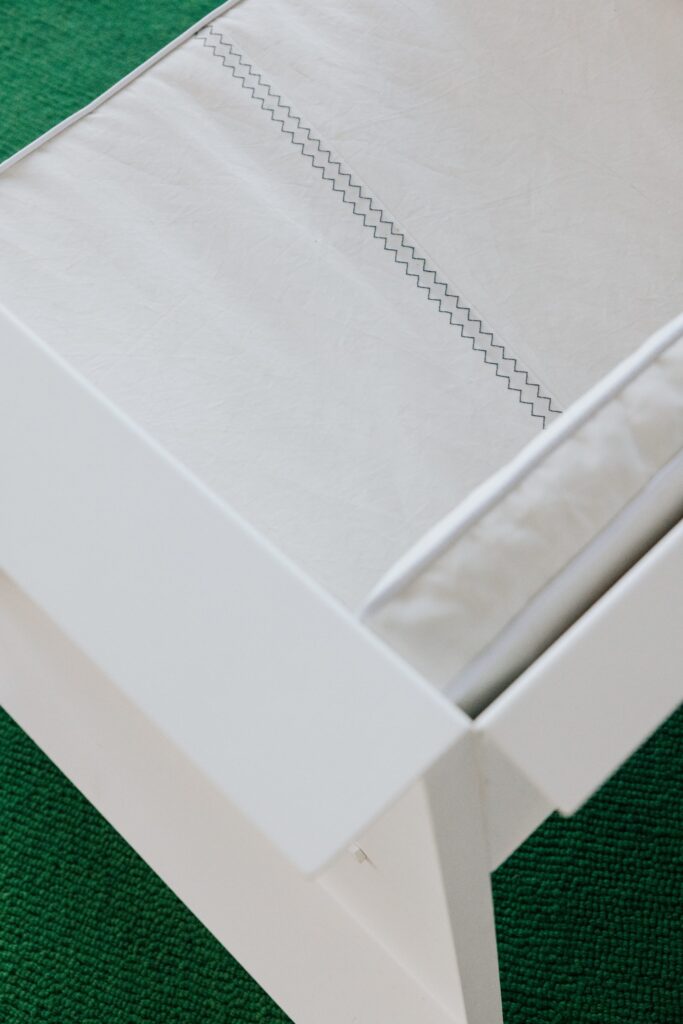 A close-up, top-down view of a minimalist white chair featuring a textured white fabric cushion with two parallel zigzag stitch lines, resting on a vibrant green textured carpet.