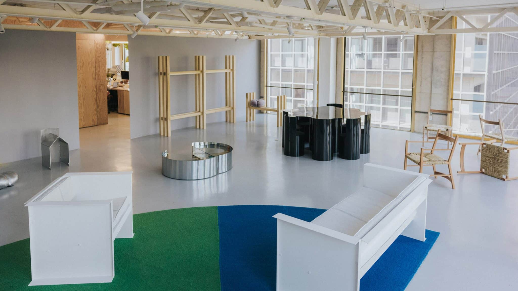 A wide-angle view of a minimalist, industrial-style gallery space featuring sculptural furniture, including white sofas on a green and blue rug, a black cylindrical table, and wooden shelving units against a grey wall.