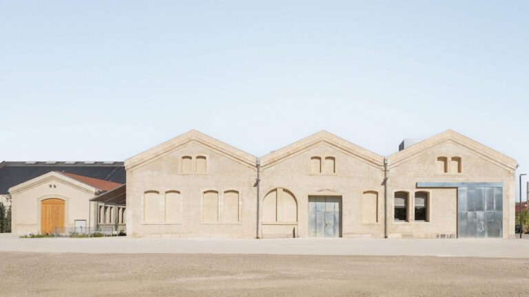 A wide, frontal view of a minimalist industrial building with three connected gabled roofs made of light-colored stone, featuring arched and rectangular windows and large metallic doors under a clear sky.