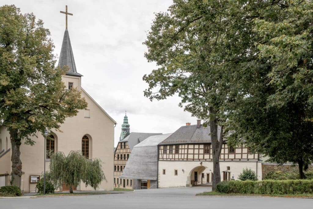 A view of a historic German square featuring a traditional church, a half-timbered building, and a modern museum addition with a sloping, grey textured roof.