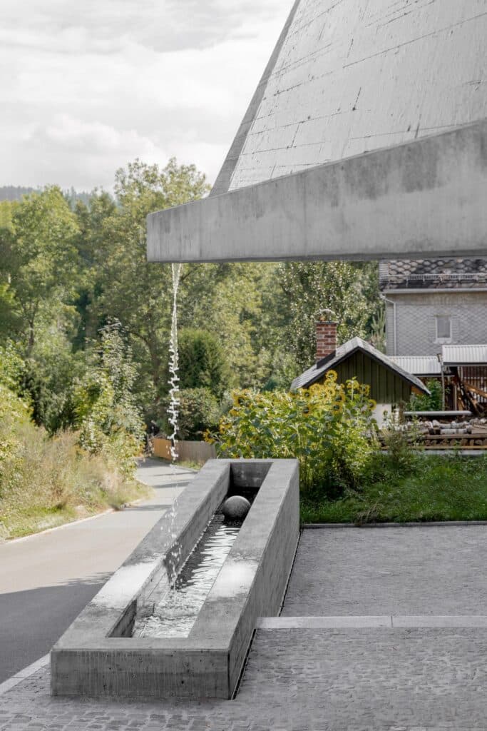 A minimalist concrete water feature where a thin stream of water falls from a cantilevered roof into a long concrete basin containing a stone sphere.