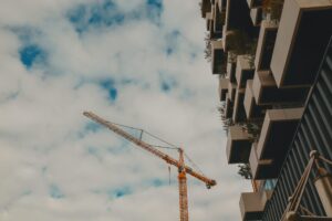 A low-angle shot of a bright yellow construction crane positioned next to a modern building featuring white, box-like staggered balconies with greenery under a cloudy sky.