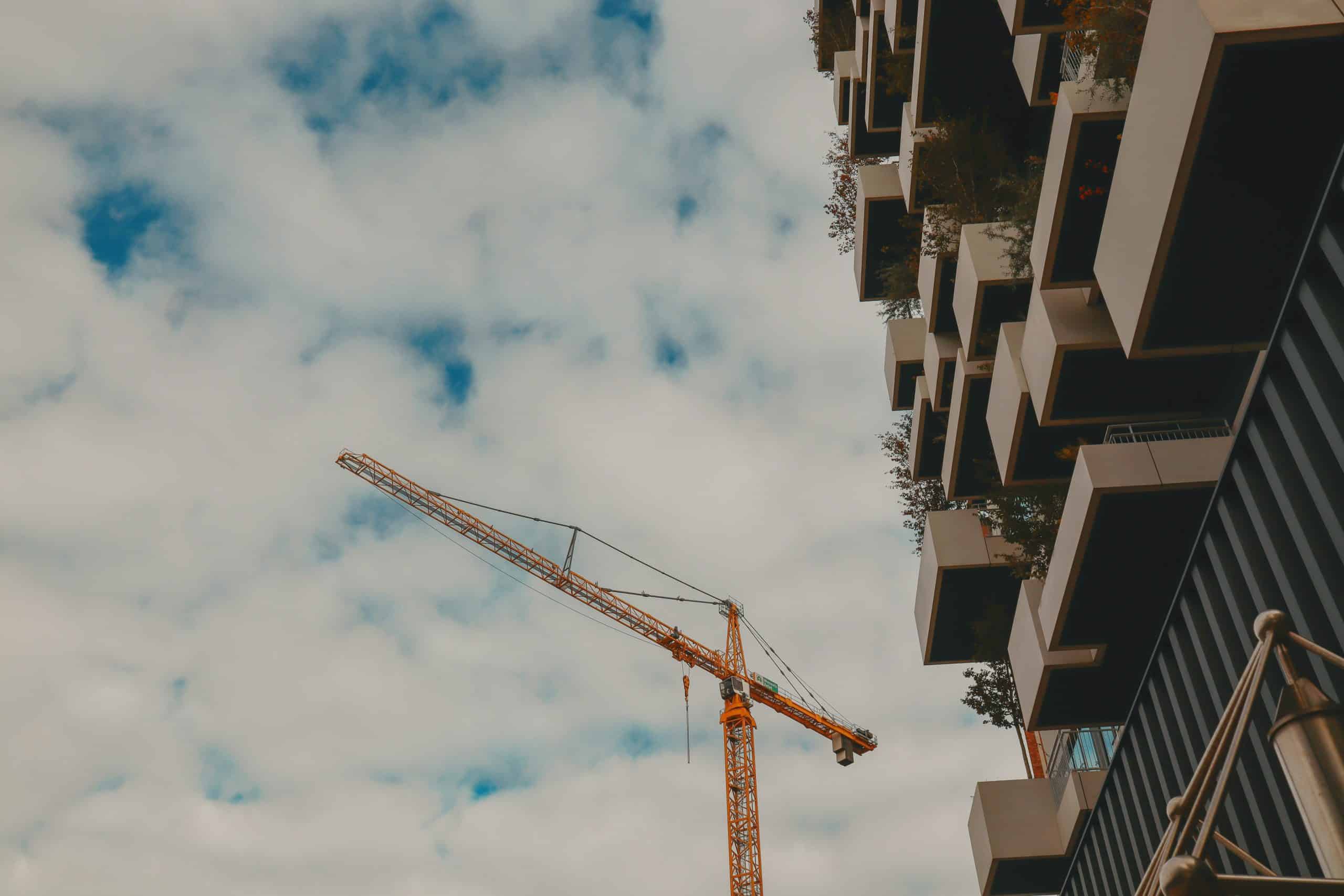 A low-angle shot of a bright yellow construction crane positioned next to a modern building featuring white, box-like staggered balconies with greenery under a cloudy sky.
