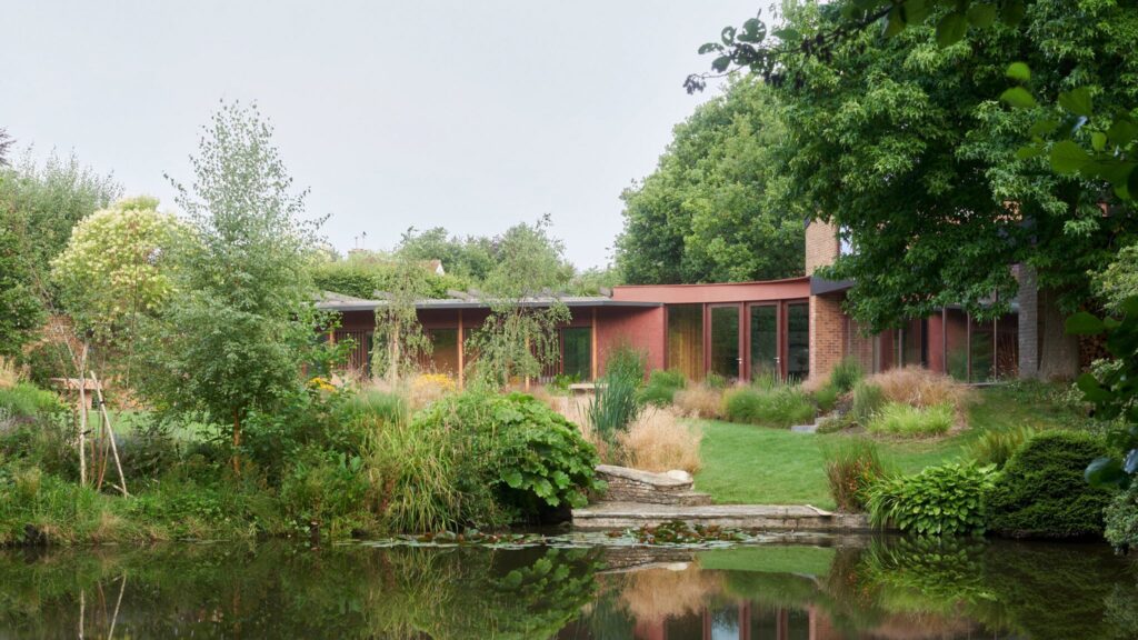 A modern curved red timber-clad residential extension overlooking a lush green garden and a reflective pond in East Sussex.