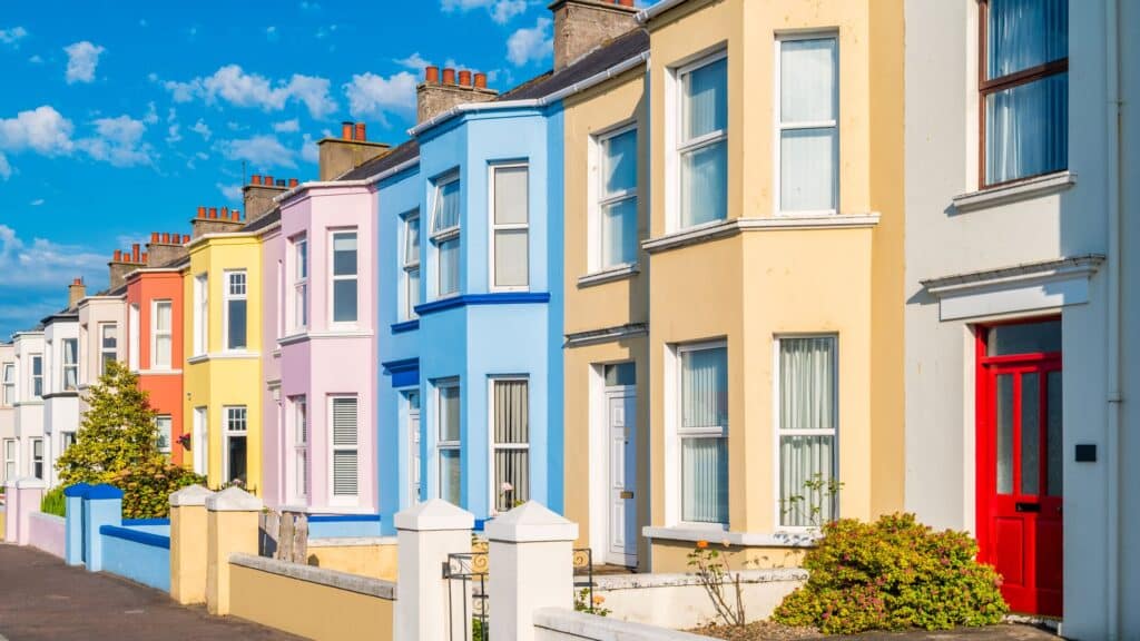A row of colorful terraced houses with bay windows under a bright blue sky. The houses are painted in pastel shades of yellow, pink, blue, and orange, featuring a vibrant red front door in the foreground.