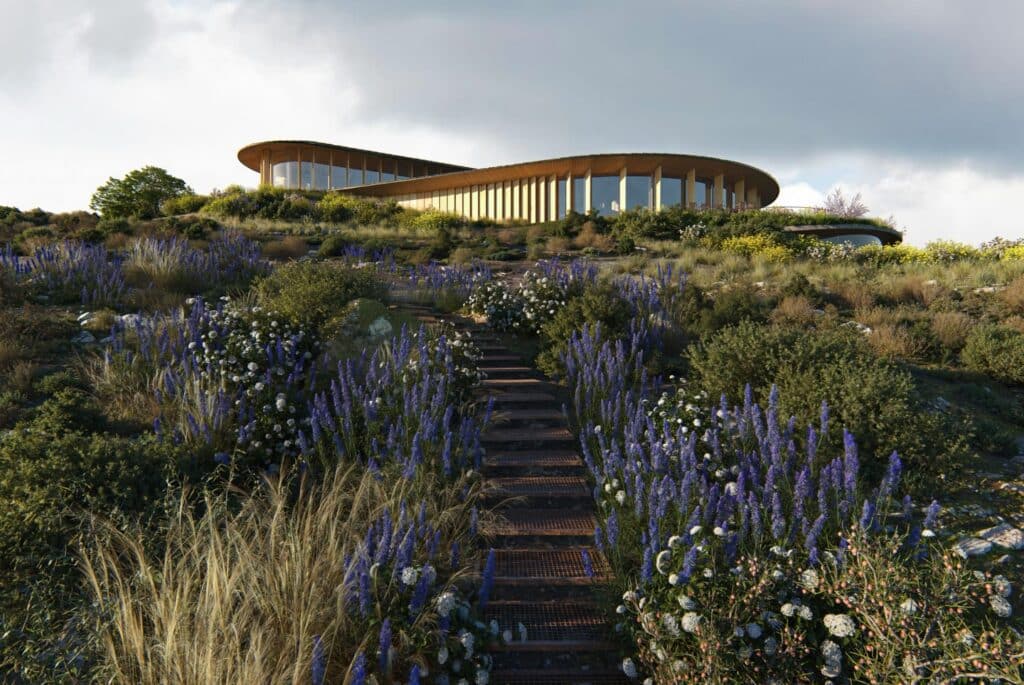 Curved mass timber pavilion sitting atop a grassy coastal hillside with a stone pathway leading up to it.
