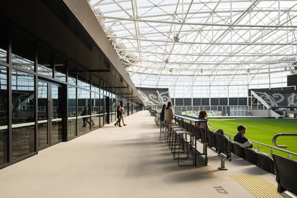 Spectators look out over the green pitch from a modern glass-enclosed concourse area