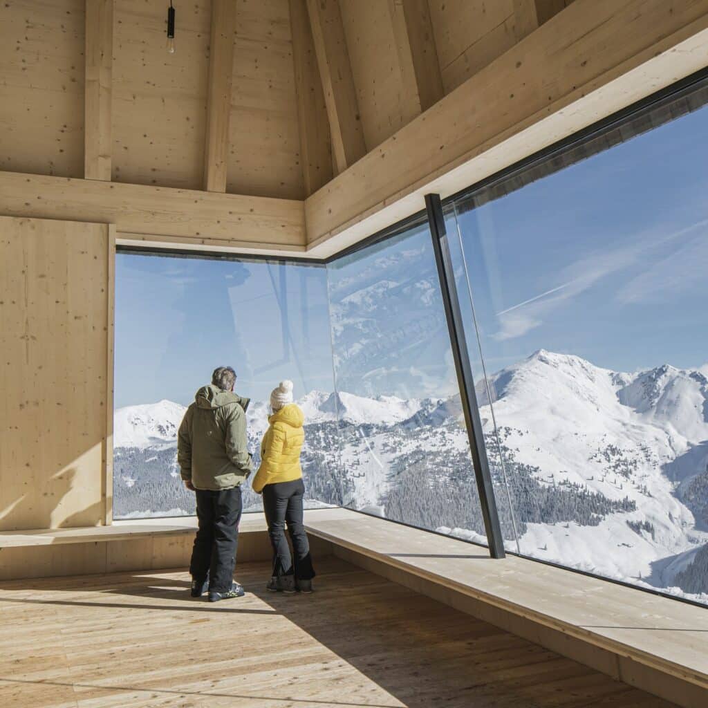 التصميم الداخلي لبرج جبال الألب يتميز بجدران خشبية فاتحة ونافذة زاوية كبيرة تعرض قمم الجبال الثلجية.