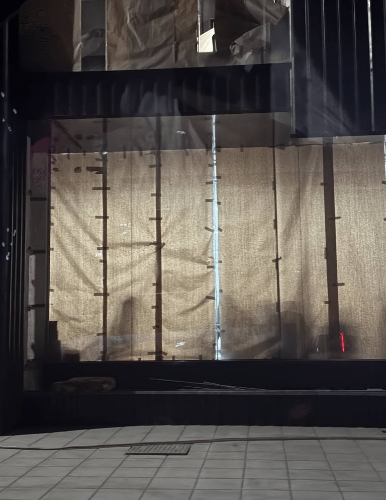 Nighttime view of a commercial storefront under construction, where brown paper taped to the glass is backlit by interior lights, accidentally revealing silhouettes of the unfinished space.