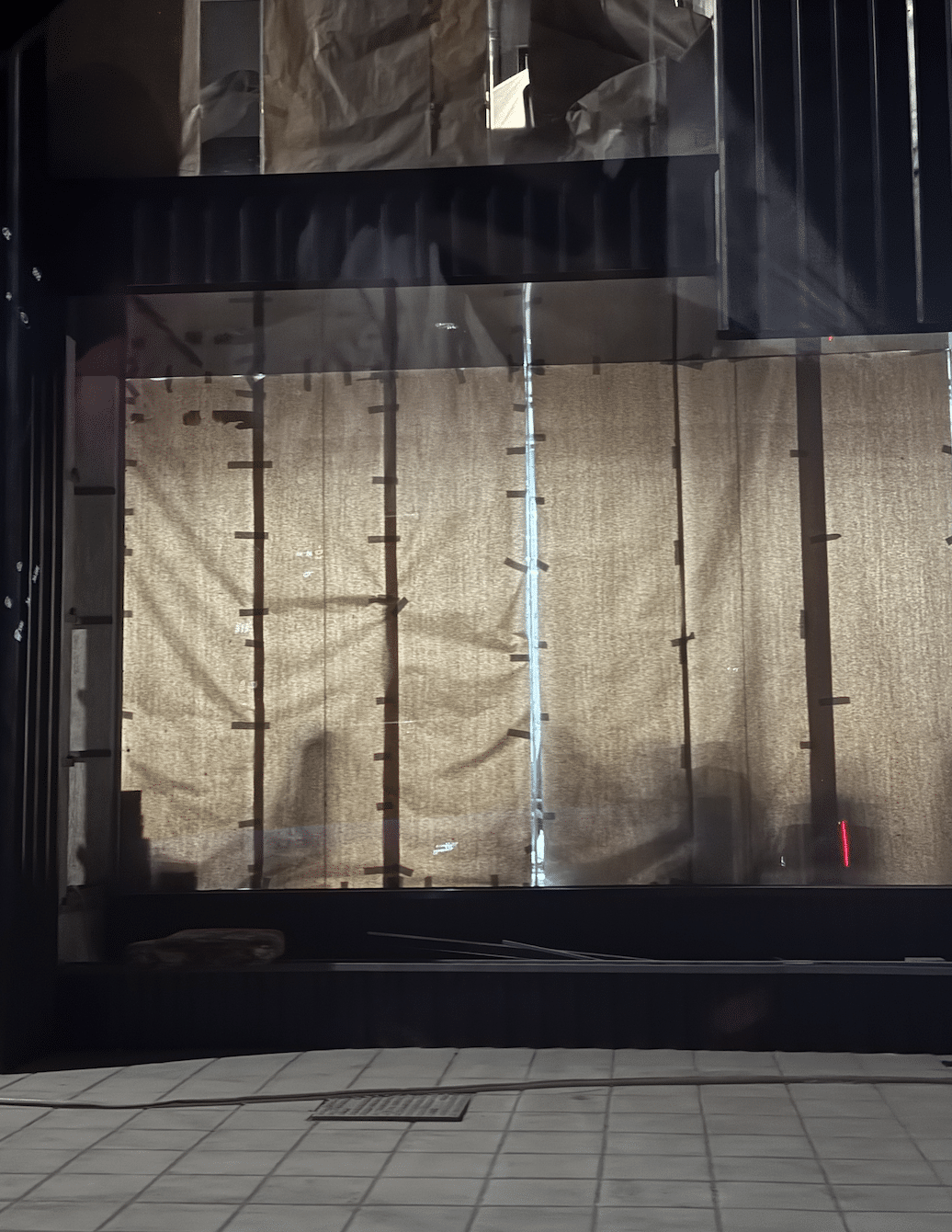 Nighttime view of a commercial storefront under construction, where brown paper taped to the glass is backlit by interior lights, accidentally revealing silhouettes of the unfinished space.