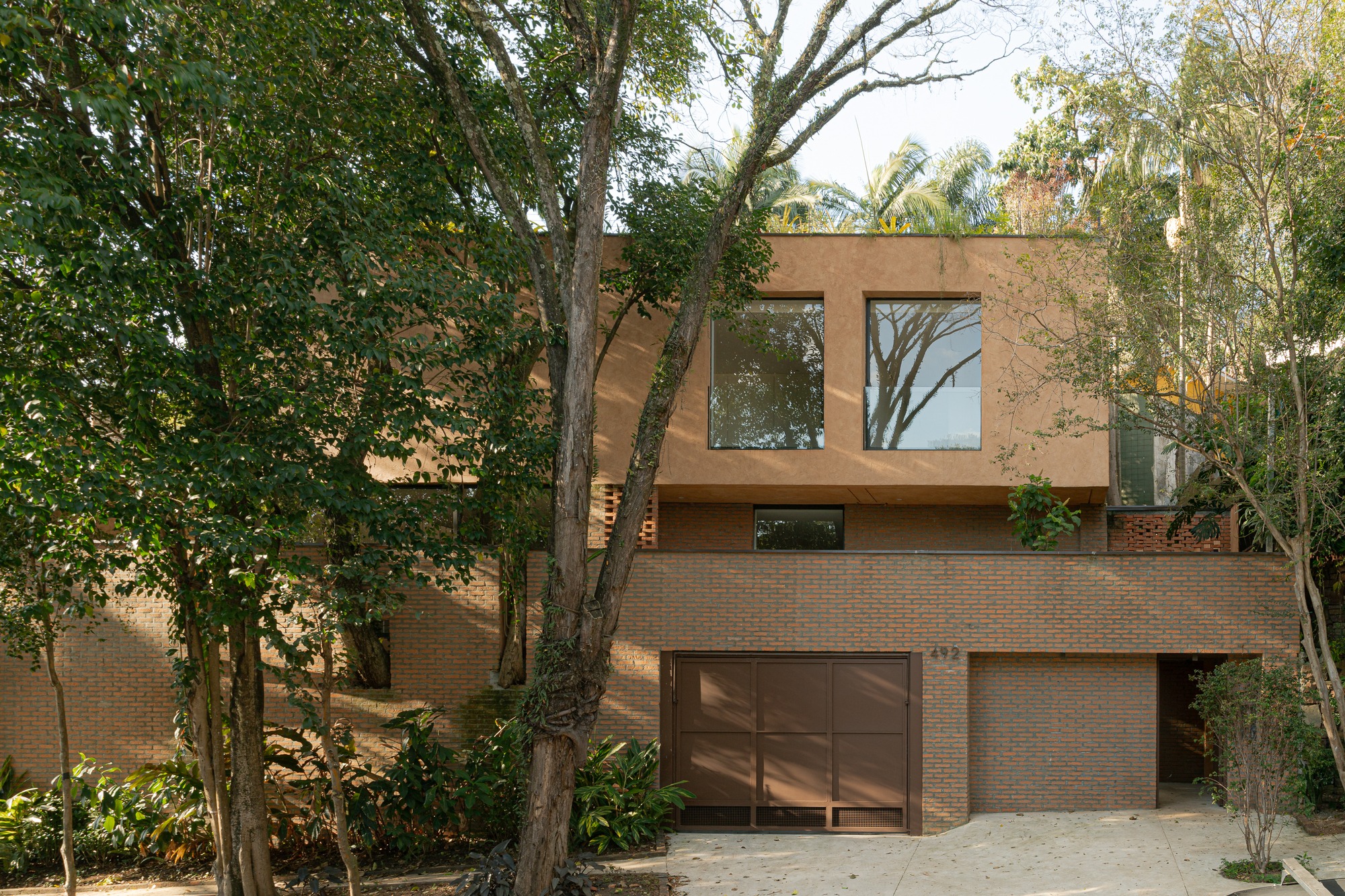 Front facade of Tamanás House showing brick ground floor and clay-colored upper volume integrated with trees.