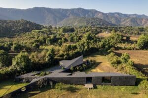 Aerial view of Casa Mavra featuring two angular black concrete volumes integrated into a lush green valley with mountain views.