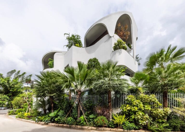 Modern white multi-story house facade with organic curved balconies and dense tropical greenery under a cloudy sky.