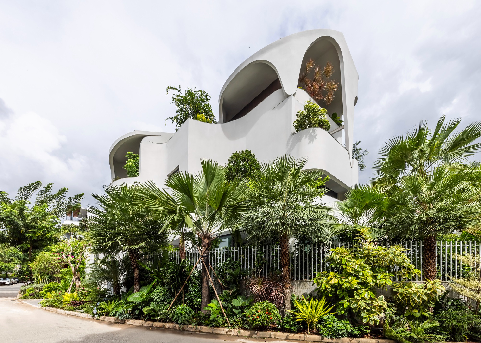 Modern white multi-story house facade with organic curved balconies and dense tropical greenery under a cloudy sky.