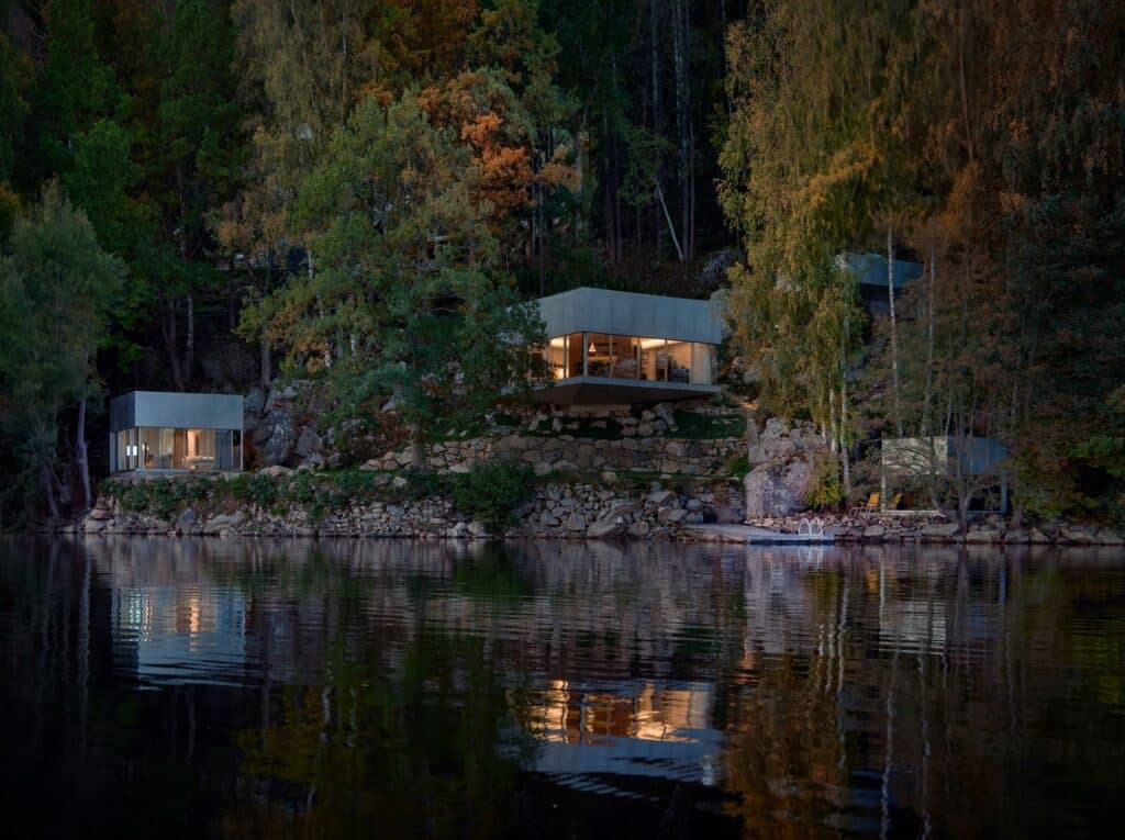 A wide dusk shot of Bor&aring;s Getaway House featuring multiple cubic volumes nestled in a steep forest slope reflecting on the lake surface.