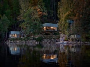A wide dusk shot of Borås Getaway House featuring multiple cubic volumes nestled in a steep forest slope reflecting on the lake surface.