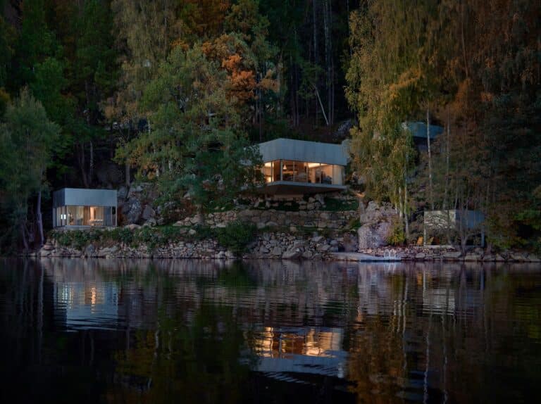 A wide dusk shot of Borås Getaway House featuring multiple cubic volumes nestled in a steep forest slope reflecting on the lake surface.
