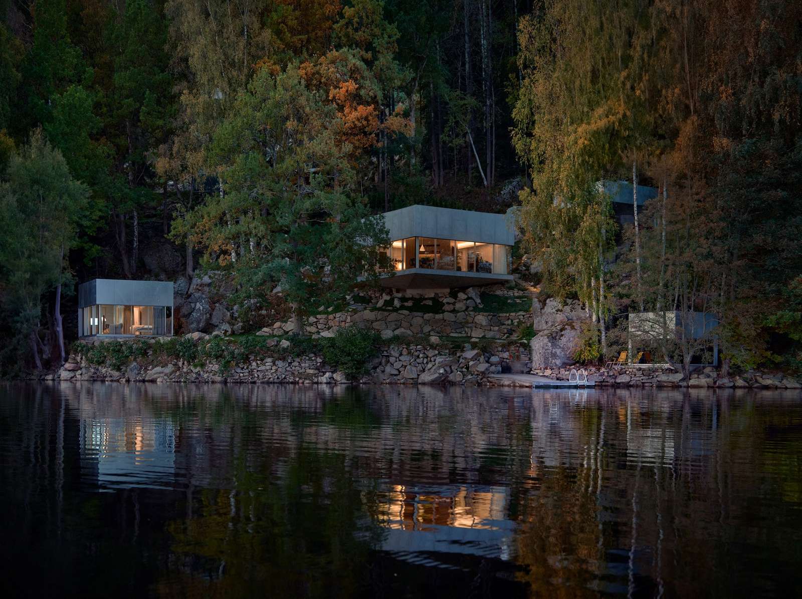 A wide dusk shot of Borås Getaway House featuring multiple cubic volumes nestled in a steep forest slope reflecting on the lake surface.