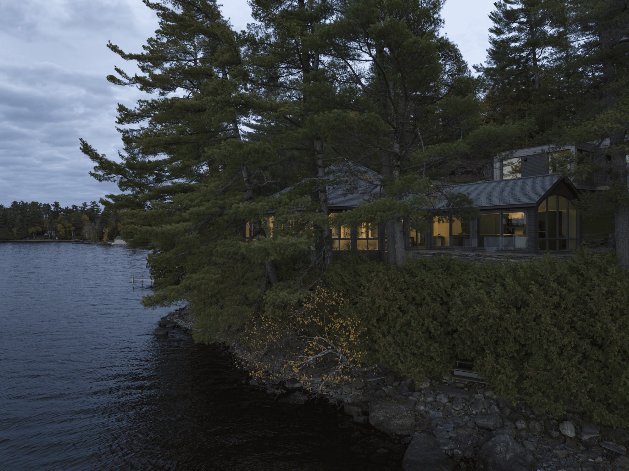 Side view of the renovated traditional cottage at Maison de la Pointe, glowing at dusk, nestled among mature pine trees on the rocky shore of Lake Memphremagog.
