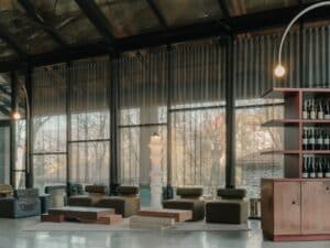 Interior lounge area with olive green seating, sheer curtains, and a copper wine display shelf against a forest view.