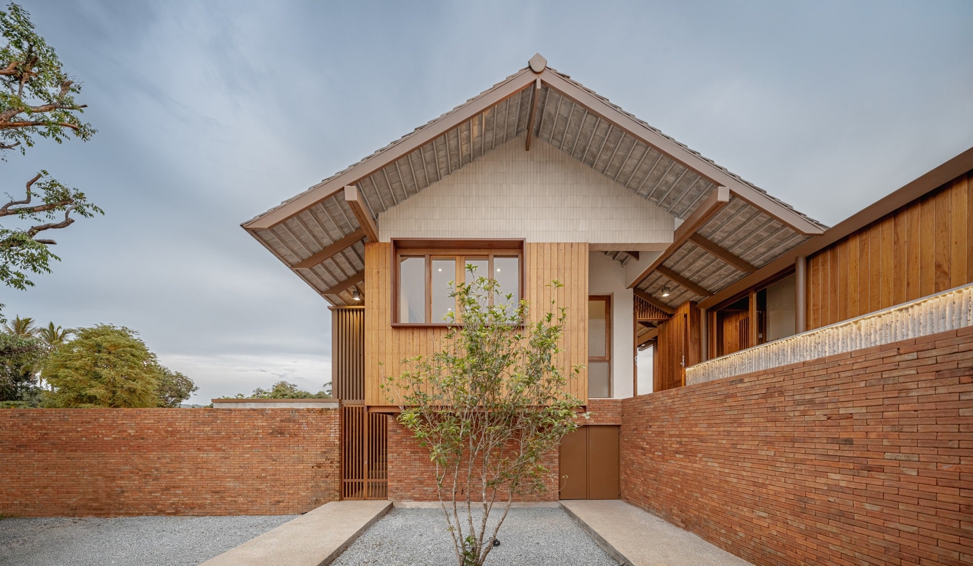 Modern Lanna house facade featuring a gabled roof, timber cladding, and a red brick boundary wall in Chiang Rai.