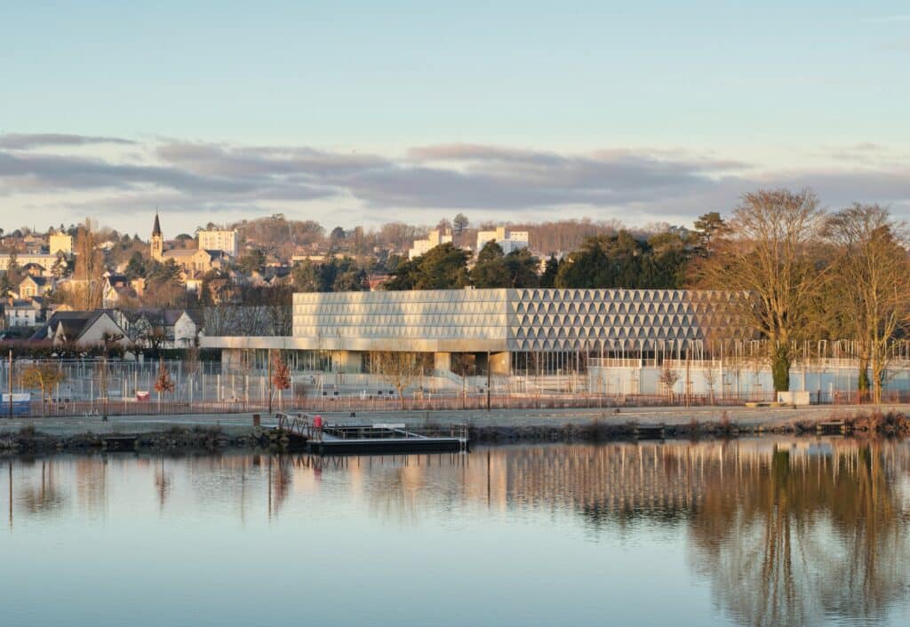 Panoramic view of the Sporting Vichy building reflected in the Allier River at sunset, showing the textured facade and surrounding trees.