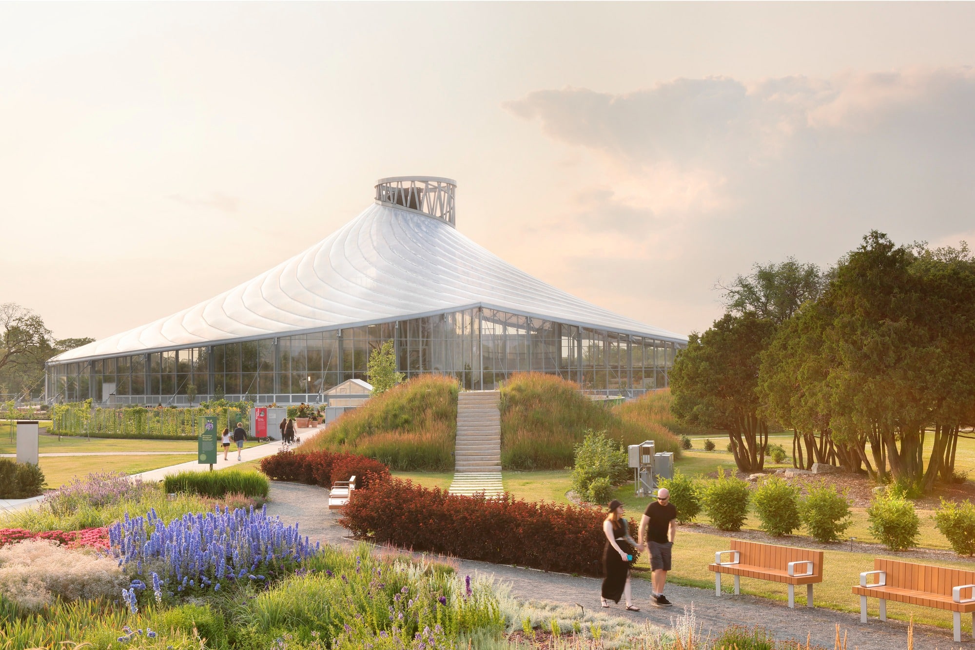 External view of The Leaf at Assiniboine Park featuring its spiral ETFE roof and glass facade surrounded by lush gardens and walking paths at sunset.