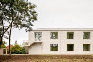Side view of Care Villa by FELT architecture showing a white brick facade with a minimalist exterior concrete staircase and large square windows under a cloudy sky.