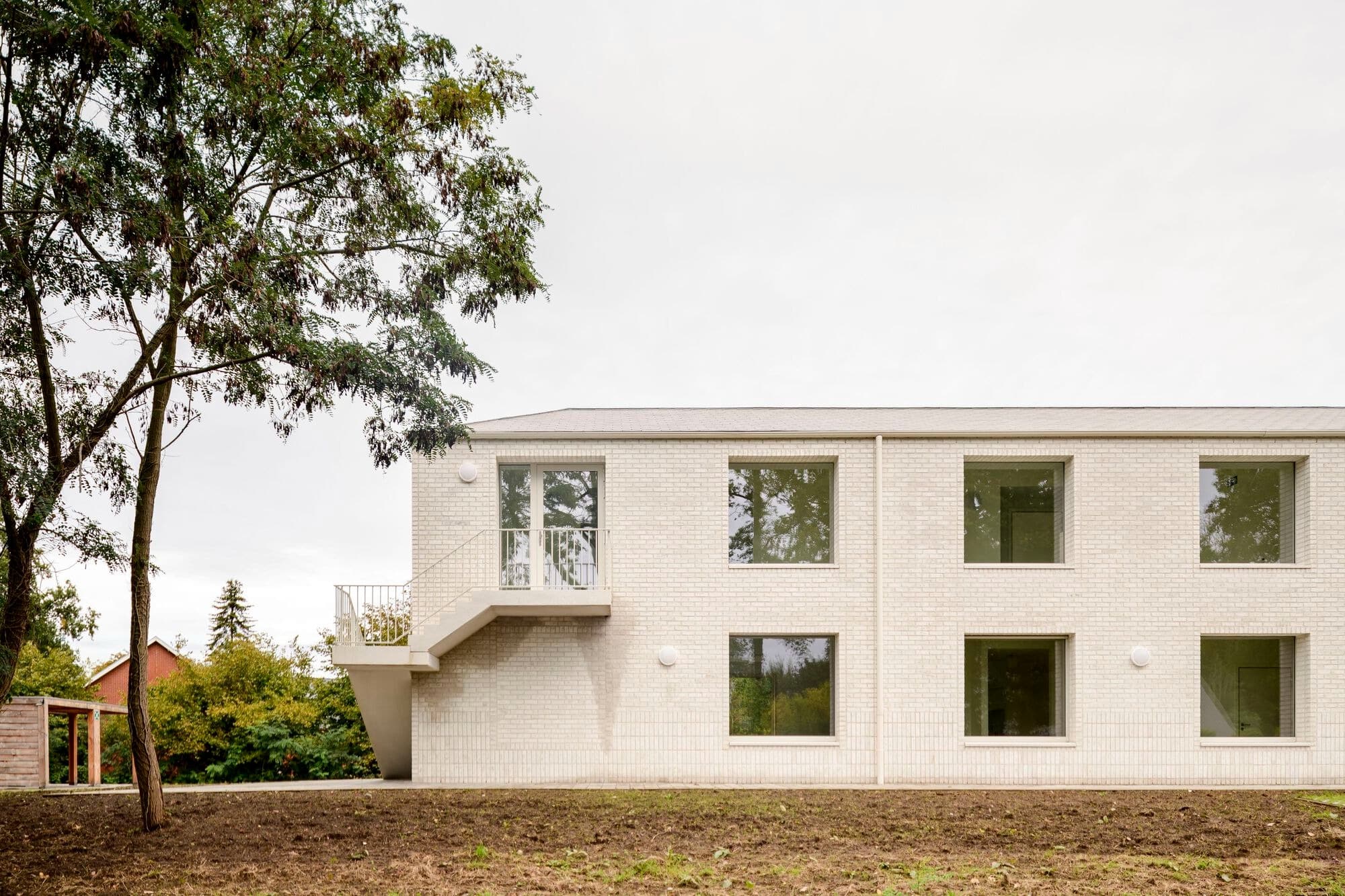 Side view of Care Villa by FELT architecture showing a white brick facade with a minimalist exterior concrete staircase and large square windows under a cloudy sky.